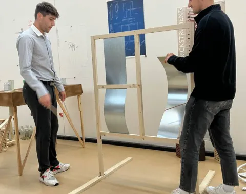 Zwei Studierende arbeiten in einem Atelier an einem Holzrahmen mit Metallteilen. Im Hintergrund sind Pläne und Modelle zu sehen.