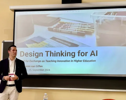 Ein Mann steht vor einer Leinwand, auf der eine Präsentation mit dem Titel "Design Thinking for AI" gezeigt wird. Der Raum wirkt wie ein Seminarraum, und es sind einige Stifte und Notizen auf der Leinwand zu sehen.