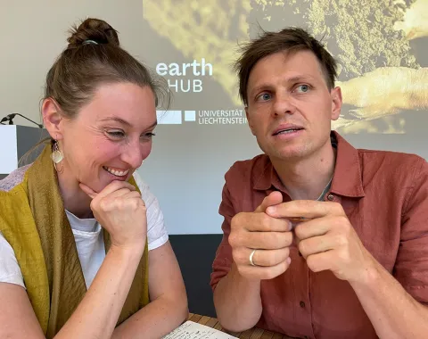 Zwei Personen sitzen an einem Tisch und diskutieren. Im Hintergrund ist ein Bildschirm mit dem Logo des Earth-Hub-Symposiums der Universität Liechtenstein zu sehen.