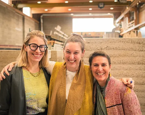 Drei lachende Personen stehen in einer Werkstatt vor Wänden aus Stampflehm. Sie tragen bunte Kleidung und umarmen sich freundschaftlich. Im Hintergrund sind Regale und Baumaterialien zu sehen.