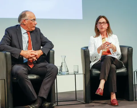 Zwei Personen sitzen auf einer Bühne in schwarzen Sesseln. Die Person links trägt einen dunklen Anzug und eine rote Krawatte, die Person rechts trägt eine weiße Jacke und rote Schuhe. Zwischen ihnen stehen Wasserkaraffen und Gläser.