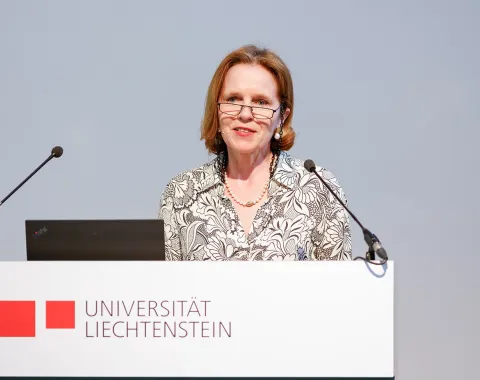 Es ist eine Frau zu sehen, welche vor einem Redepult mit dem Logo der Universität Liechtenstein steht.