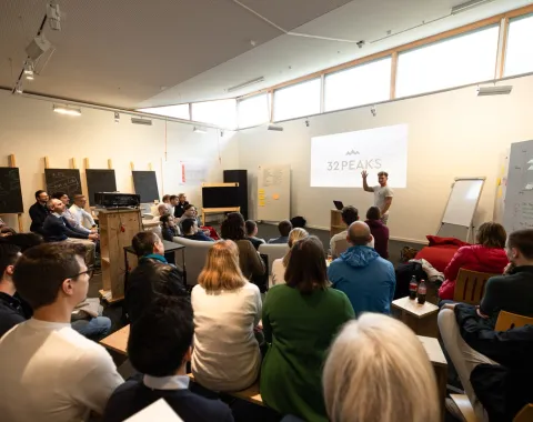 Eine Gruppe von Menschen sitzt in einem modernen Seminarraum und hört einem Vortragenden zu, der vor einer Leinwand mit der Aufschrift "32PEAKS" steht. Der Raum ist hell und mit großen Fenstern ausgestattet.