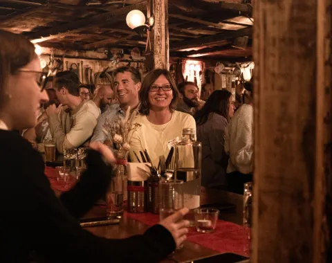 Eine Gruppe von Menschen sitzt in einer gemütlichen, rustikalen Bar. Im Vordergrund unterhalten sich Personen an einem Tisch, während im Hintergrund weitere Gäste lächeln und die Atmosphäre genießen. Die Szene wirkt lebhaft und einladend.