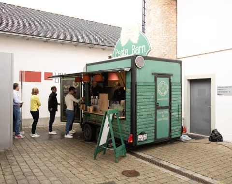 Ein grüner Foodtruck mit der Aufschrift "Pasta Barn" steht auf einem gepflasterten Platz. Mehrere Personen stehen in einer Schlange vor dem Truck und warten auf ihre Bestellung. Im Hintergrund sind Gebäude der Universität Liechtenstein zu sehen.