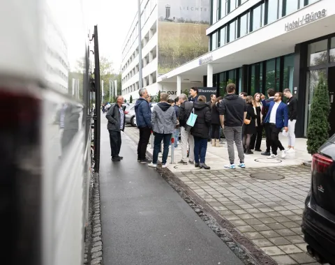 Gruppe von Menschen steht vor einem modernen Gebäude auf dem Campus der Universität Liechtenstein, möglicherweise während einer Veranstaltung. Im Vordergrund sind geparkte Autos zu sehen.
