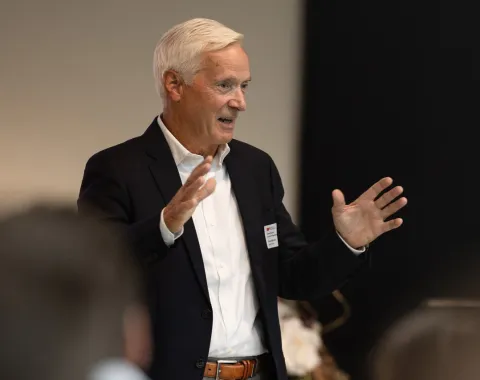 Ein älterer Mann in einem dunklen Anzug spricht gestikulierend bei einer Veranstaltung. Im Hintergrund sind unscharfe Personen zu erkennen.