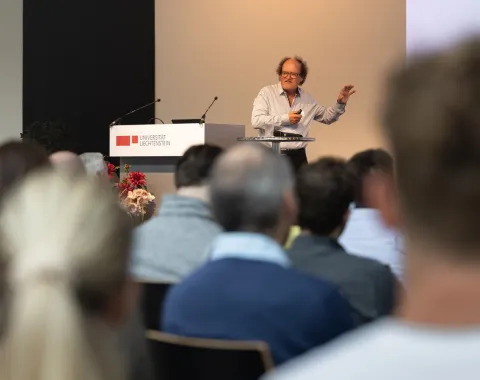 Ein Redner steht an einem Podium und spricht zu einem Publikum bei einer Veranstaltung an der Universität Liechtenstein. Im Hintergrund ist das Logo der Universität sichtbar.