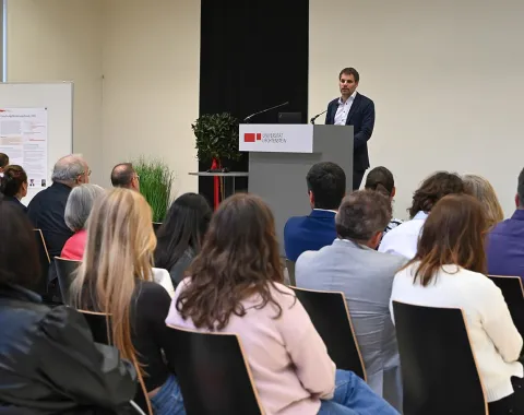Rektor Christian Frommelt hält eine Rede am Rednerpult vor einem sitzenden Publikum in einem Konferenzraum. Im Hintergrund ist ein Poster sichtbar.