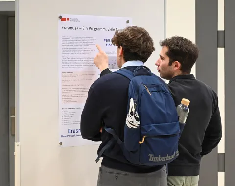 Zwei Personen betrachten ein Poster über das Erasmus-Programm an einer Wand. Eine Person zeigt auf den Text. Beide tragen Freizeitkleidung, einer hat einen Rucksack.