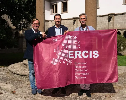 Drei Personen halten ein Banner mit der Aufschrift "ERCIS - European Research Center for Information Systems" in einem sonnigen Innenhof. Im Hintergrund sind historische Gebäude und Säulen zu sehen.