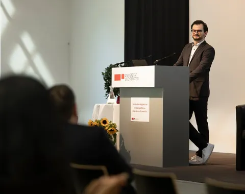 Ein Mann steht an einem Rednerpult in einem Konferenzraum der Universität Liechtenstein. Er trägt einen Anzug und spricht zu einem Publikum. Im Hintergrund sind Sonnenblumen und ein Fenster zu sehen.
