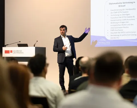 Ein Mann hält einen Vortrag vor einem Publikum an der Universität Liechtenstein. Er steht neben einer Leinwand mit einer Präsentation. Im Vordergrund sind die Köpfe der Zuhörer zu sehen.