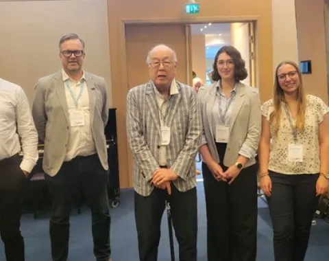 Fünf Personen stehen nebeneinander in einem Konferenzraum. Sie tragen Namensschilder und lächeln in die Kamera. Im Hintergrund sind Türen und ein Tisch sichtbar.