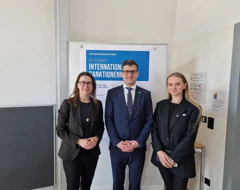 Drei Personen stehen vor einem Poster mit der Aufschrift "Intensivkurs Internationales Sanktionenrecht". Sie tragen formelle Kleidung und lächeln in die Kamera.