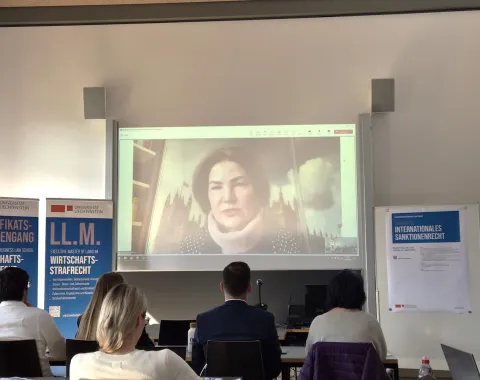 Vortrag im Rahmen des Intensivkurses „Internationales Sanktionenrecht“ an der Universität Liechtenstein. Eine Rednerin wird per Videokonferenz auf eine Leinwand projiziert. Im Vordergrund sitzen Teilnehmende, im Hintergrund sind Banner der Universität zu sehen.