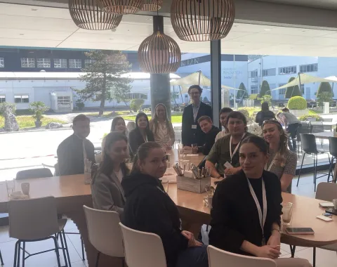 Studierende der Universität Liechtenstein sitzen in einer modernen Cafeteria bei Ivoclar in Schaan. Sie sind um einen Tisch versammelt und blicken in die Kamera. Im Hintergrund sind große Fenster mit Blick auf den Außenbereich zu sehen.