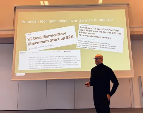 Ein Vortragender steht vor einer Präsentationsfolie, die Schlagzeilen über die Übernahme des deutschen KI-Start-ups G2K durch den amerikanischen Technologiekonzern ServiceNow zeigt.