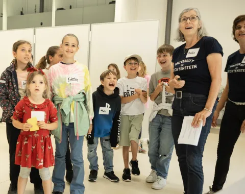 Eine Gruppe von Kindern und zwei Erwachsenen steht in einem hellen Raum. Die Kinder wirken fröhlich und interessiert. Die Erwachsenen tragen T-Shirts mit dem Logo der Universität Liechtenstein.