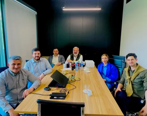 Sechs Personen sitzen an einem Besprechungstisch in einem modernen Konferenzraum und nehmen an einem Meeting oder Workshop teil.