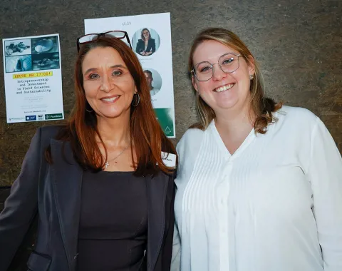 Zwei lächelnde Personen stehen vor einer Wand mit mehreren Postern. Eine Person trägt einen Blazer, die andere ein weißes Hemd.