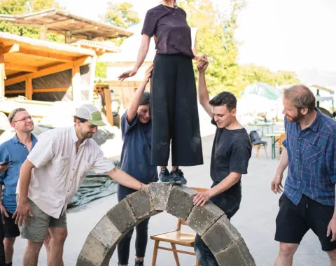 Ein Gruppe von Menschen baut einen selbsttragenden Lehmbogen im Freien. Eine Person steht oben auf dem Bogen, während andere sie stützen. Im Hintergrund sind Bäume und Gebäude zu sehen.