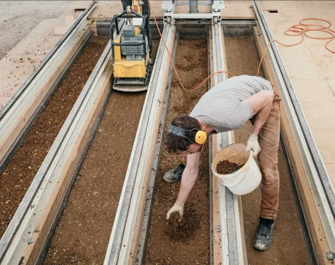 Eine Person arbeitet an einer Lehmbau-Konstruktion, indem sie Erde in eine Form füllt. Im Hintergrund ist eine Maschine zu sehen, die möglicherweise zur Verdichtung des Materials verwendet wird. Die Szene zeigt praktische Anwendung von Lehmbau-Techniken im EARTH HUB der Universität Liechtenstein.