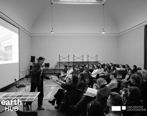 Ein Vortragender steht vor einem Publikum in einem großen Raum mit hoher Decke. Auf der Leinwand ist eine Projektion zu sehen. Im Vordergrund befindet sich ein Tisch mit Materialien. Das Logo der Universität Liechtenstein und des earth HUB ist sichtbar.