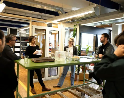 Personen stehen um einen grünen Tisch in einem modernen Raum mit Glaswänden und Pflanzen. Eine Person hält ein Papier und spricht zu den anderen. Die Szene wirkt wie ein informelles Treffen oder eine Diskussion.