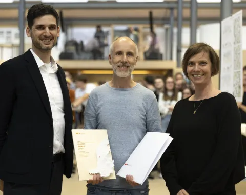 Drei Personen stehen lächelnd in einem Innenraum. Eine Person hält ein Zertifikat in der Hand. Im Hintergrund sind unscharf weitere Menschen zu sehen.
