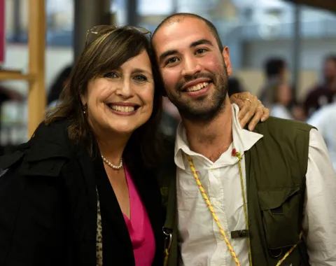 Zwei Personen lächeln in die Kamera bei einer Veranstaltung. Die Frau trägt eine Perlenkette und ein pinkes Oberteil, der Mann ein weißes Hemd. Sie stehen nah beieinander in einem Innenraum.