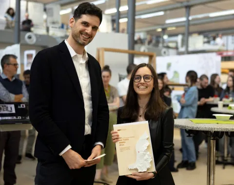 Zwei Personen stehen lächelnd in einem modernen Innenraum während einer Veranstaltung. Eine Person hält ein Buch mit einer Karte auf dem Cover. Im Hintergrund sind weitere Personen und Ausstellungstafeln zu sehen.