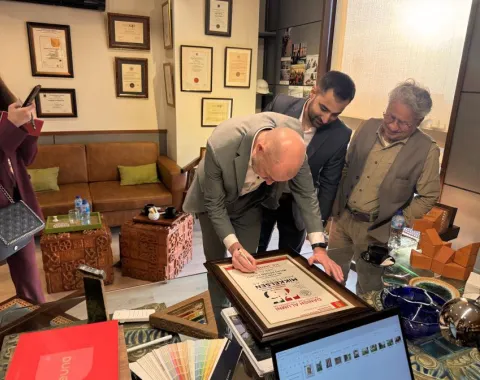 Ein Mann in einem Anzug unterschreibt eine Auszeichnung in einem Büro, während zwei weitere Männer zuschauen. Im Hintergrund sind gerahmte Zertifikate an der Wand zu sehen. Auf dem Tisch liegen Farbmuster und ein Laptop.