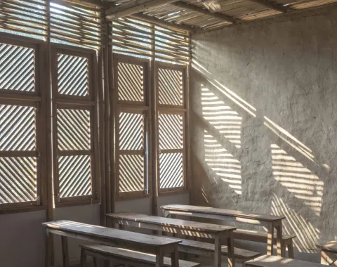 Innenraum eines nachhaltig gebauten Klassenzimmers mit Holzbänken und Wänden aus natürlichen Materialien. Sonnenlicht fällt durch hölzerne Fensterläden und erzeugt Muster auf der Wand.