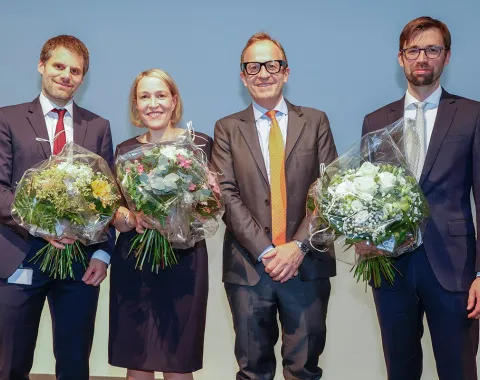 Vier Personen stehen nebeneinander auf einer Bühne. Drei von ihnen halten Blumensträuße in den Händen. Alle tragen formelle Kleidung und lächeln in die Kamera.