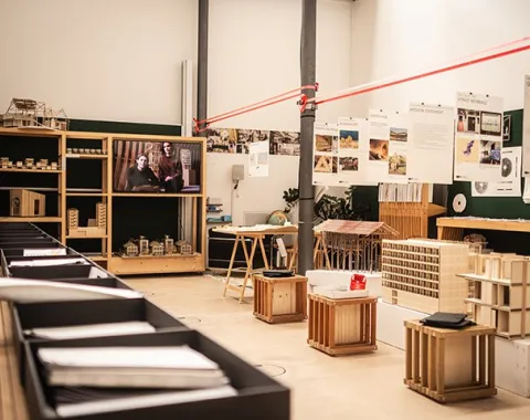 Ein Raum der Liechtenstein School of Architecture mit Ausstellungsstücken und Modellen. Im Vordergrund sind Tische mit Architekturmodellen, im Hintergrund ein Bildschirm mit einer Präsentation. An den Wänden hängen Plakate mit architektonischen Entwürfen.