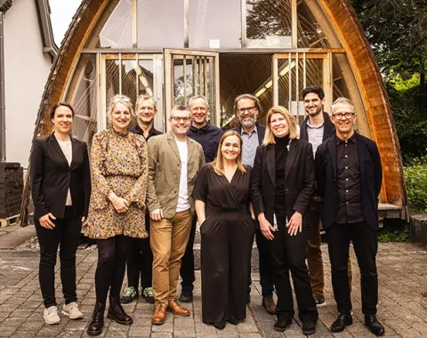 Eine Gruppe von zehn Personen steht lächelnd vor einem modernen Holzgebäude mit großen Fenstern. Die Szene vermittelt eine freundliche und professionelle Atmosphäre.