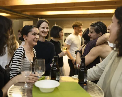 Eine Gruppe von Menschen steht um einen Tisch und unterhält sich fröhlich bei einem Event. Einige Personen halten Getränke in der Hand, während andere sich umarmen. Die Atmosphäre wirkt lebhaft und gesellig.