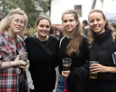 Vier junge Frauen stehen lächelnd zusammen auf einem Sommerfest der Universität Liechtenstein. Sie halten Getränke in der Hand und sind von anderen Feiernden umgeben. Im Hintergrund sind Bäume und ein Gebäude zu sehen.