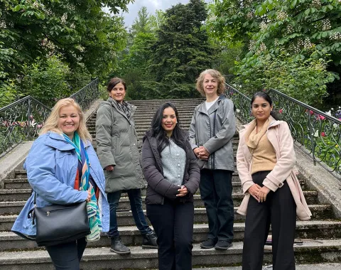 Fünf Personen stehen auf einer Treppe im Freien, umgeben von grünen Bäumen. Sie lächeln in die Kamera.