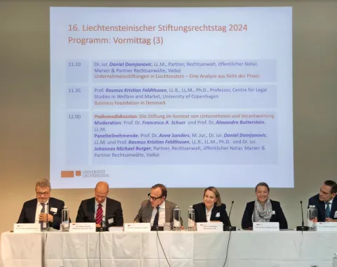 Podiumsdiskussion beim 16. Liechtensteinischen Stiftungsrechtstag 2024 an der Universität Liechtenstein. Fünf Expertinnen und Experten sitzen an einem Tisch vor einer Leinwand mit dem Tagesprogramm.