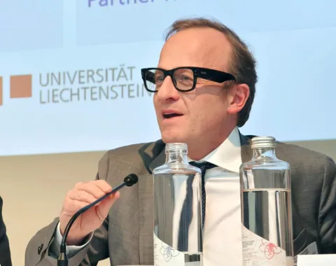 Ein Mann in Anzug und Brille spricht in ein Mikrofon auf einer Konferenz im Auditorium der Universität Liechtenstein. Vor ihm stehen Wasserflaschen. Im Hintergrund ist das Logo der Universität zu sehen.