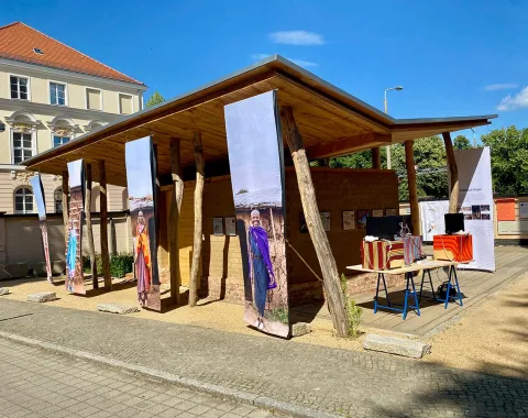 Ein hölzerner Pavillon mit großen, vertikalen Bannern, die Maasai-Frauen in traditioneller Kleidung zeigen. Im Vordergrund stehen Tische mit bunten Stoffen und Modellen. Der Pavillon ist Teil einer Ausstellung über Maasai-Baumeisterinnen und nachhaltige Architektur.