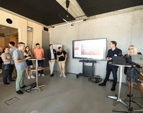 Eine Gruppe von Menschen steht in einem modernen Raum mit Betonwänden. Im Vordergrund sind Stehtische und ein großer Bildschirm an der Wand, auf dem eine Präsentation gezeigt wird. Eine Person spricht zu den Anwesenden, während andere aufmerksam zuhören. Der Raum wirkt hell und einladend.