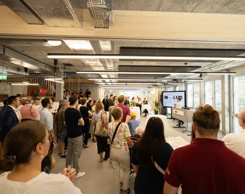Besuchergruppe beim Tag der offenen Tür an der Universität Liechtenstein, neuer Standort Ebaholz. Menschen stehen in einem modernen Raum und hören einer Präsentation zu. Im Hintergrund sind große Fenster und eine Leinwand zu sehen.