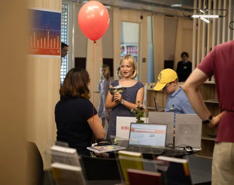 Eine Gruppe von Menschen steht an einem Informationsstand in einem modernen Gebäude. Eine Frau sitzt hinter dem Tisch, während ein Mädchen mit einem roten Ballon in der Hand lächelt. Ein Junge mit gelber Kappe betrachtet die ausgestellten Materialien. Im Hintergrund sind Regale und weitere Personen zu sehen.