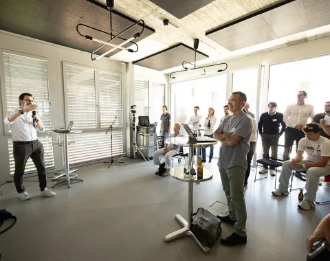 Eine Gruppe von Menschen steht in einem modernen, hellen Raum. Ein Mann hält eine Präsentation vor einem Publikum, das aufmerksam zuhört. Der Raum hat große Fenster und moderne Beleuchtung.