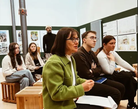 Eine Gruppe von Studierenden und Dozenten sitzt in einem Seminarraum und hört aufmerksam zu. Im Hintergrund sind Präsentationstafeln mit städtischen Entwürfen zu sehen.
