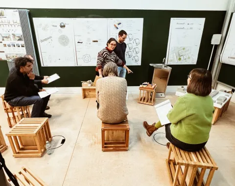 Eine Gruppe von sechs Personen sitzt auf Holzkisten in einem Raum mit Plakaten an der Wand. Zwei Personen stehen und diskutieren, während die anderen Notizen machen. Der Raum wirkt wie ein Workshop oder eine Präsentation.