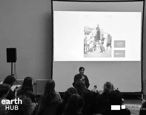 Eine Vortragende steht vor einem Publikum und präsentiert. Im Hintergrund ist eine Leinwand zu sehen. Das Bild ist in schwarz-weiss.